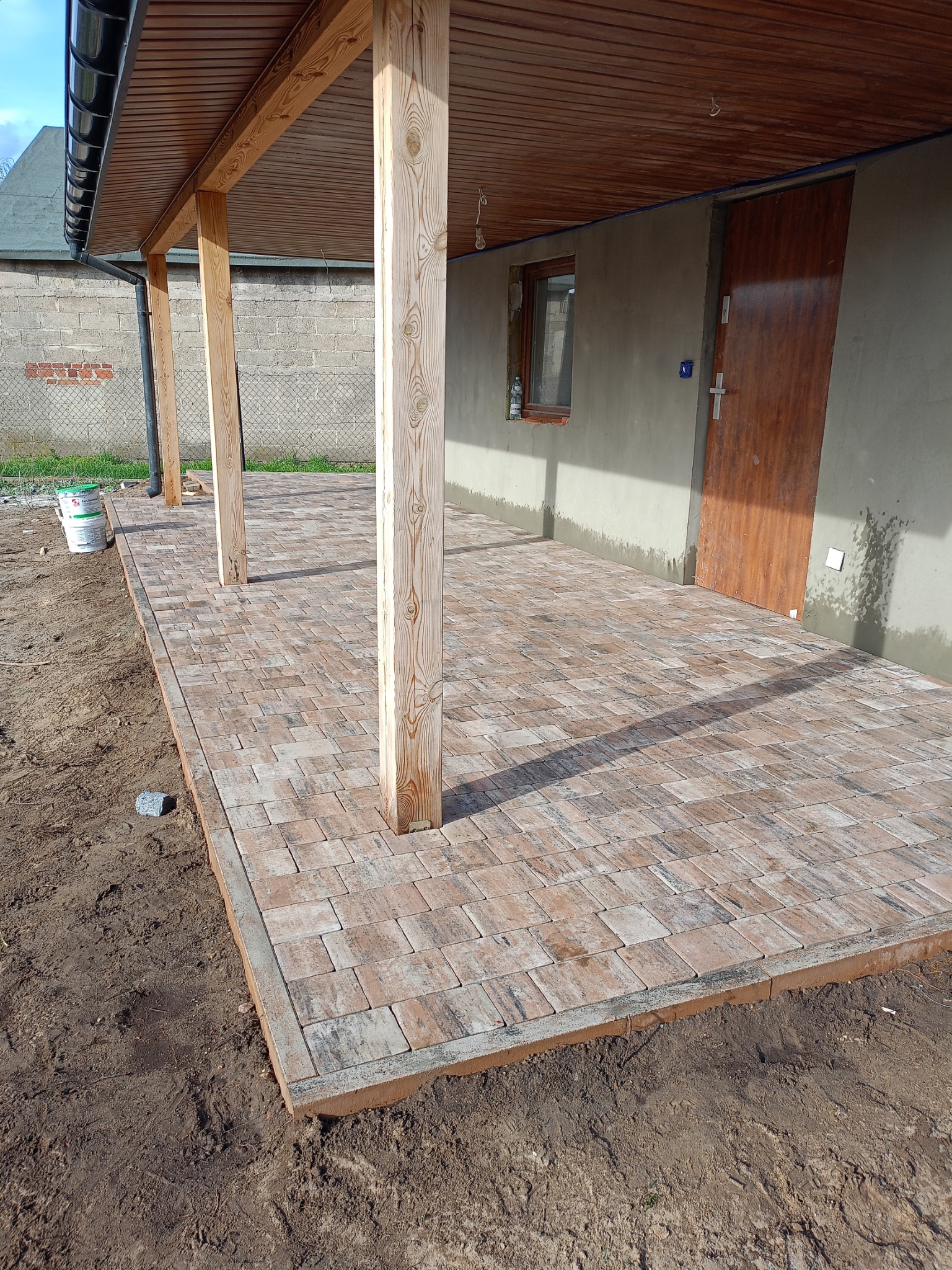 Zadaszony taras wyłożony kostką brukową w odcieniach brązu i beżu, z widocznymi drewnianymi słupami nośnymi i fragmentem elewacji budynku w surowym tynku oraz z puszkami farb w tle.