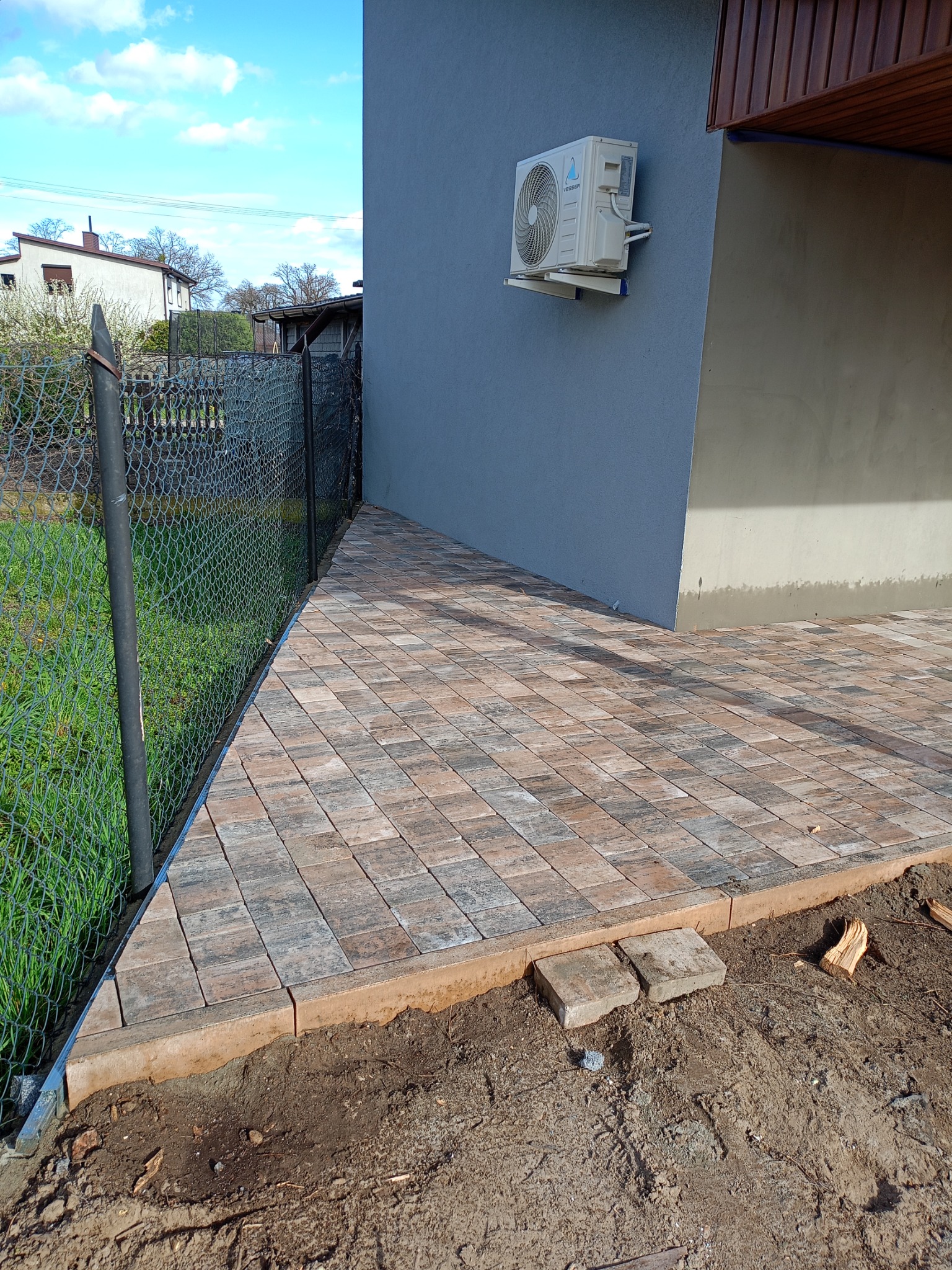 Ułożona kostka brukowa tworząca ścieżkę przy szarej ścianie budynku z zamontowaną klimatyzacją, widoczna siatka ogrodzeniowa i fragment trawnika.
