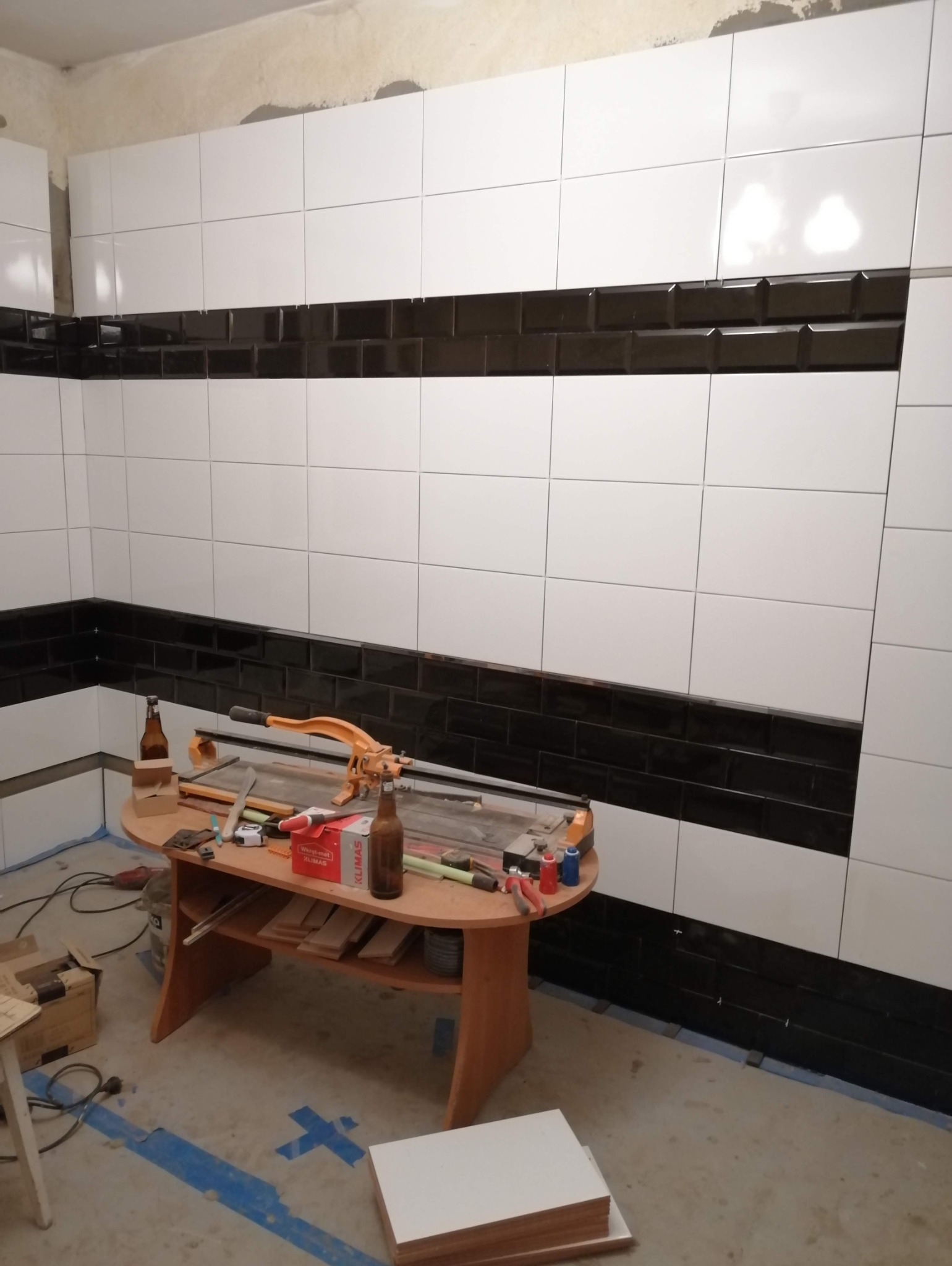 W trakcie układania białych i czarnych płytek ściennych w pomieszczeniu, widoczny stół z narzędziami do cięcia płytek i materiałami budowlanymi oraz butelki po piwie.