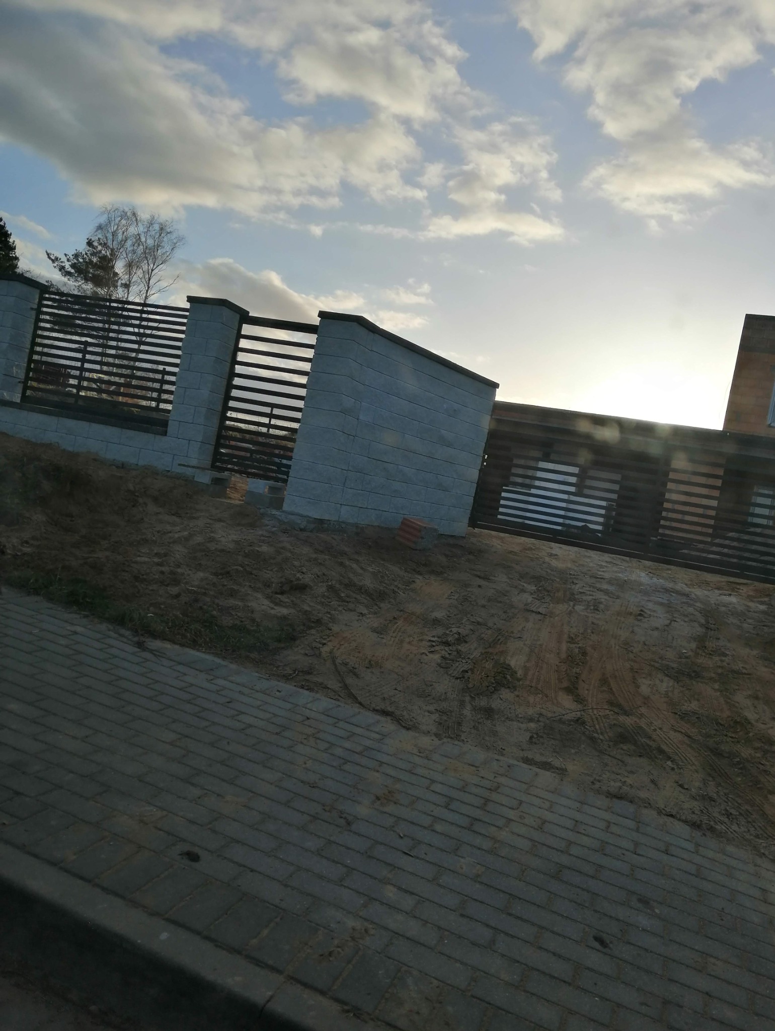 Nowoczesna, częściowo ukończona elewacja z szarej cegły i drewnianych paneli, widoczna w pochmurny dzień z brukowanym chodnikiem na pierwszym planie i ziemią w tle.