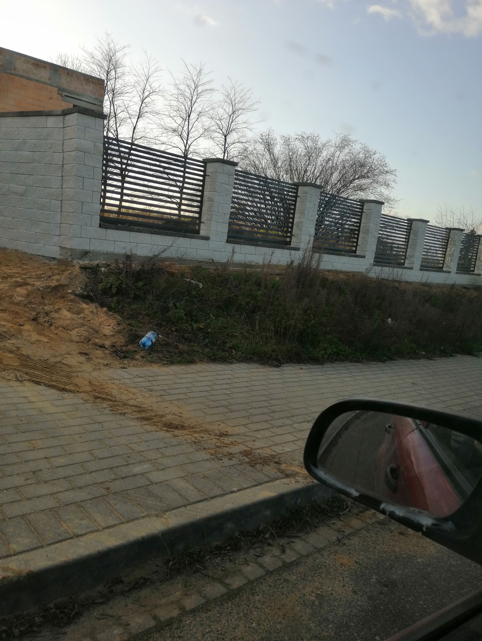 Nowoczesne ogrodzenie panelowe z betonowymi słupkami, widok zza szyby samochodu, na pierwszym planie kostka brukowa i butelka plastikowa na ziemi.
