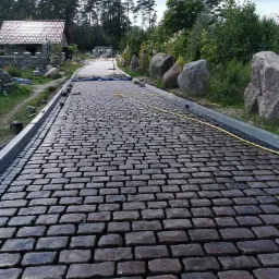 Ułożona, mokra kostka granitowa wzdłuż ścieżki z szarymi obrzeżami, widok z perspektywy, w tle budynek z czerwoną dachówką i drzewa.