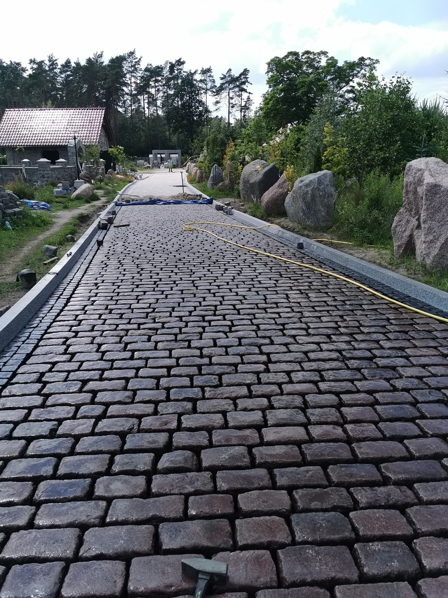 Ułożona, mokra kostka granitowa wzdłuż ścieżki z szarymi obrzeżami, widok z perspektywy, w tle budynek z czerwoną dachówką i drzewa.
