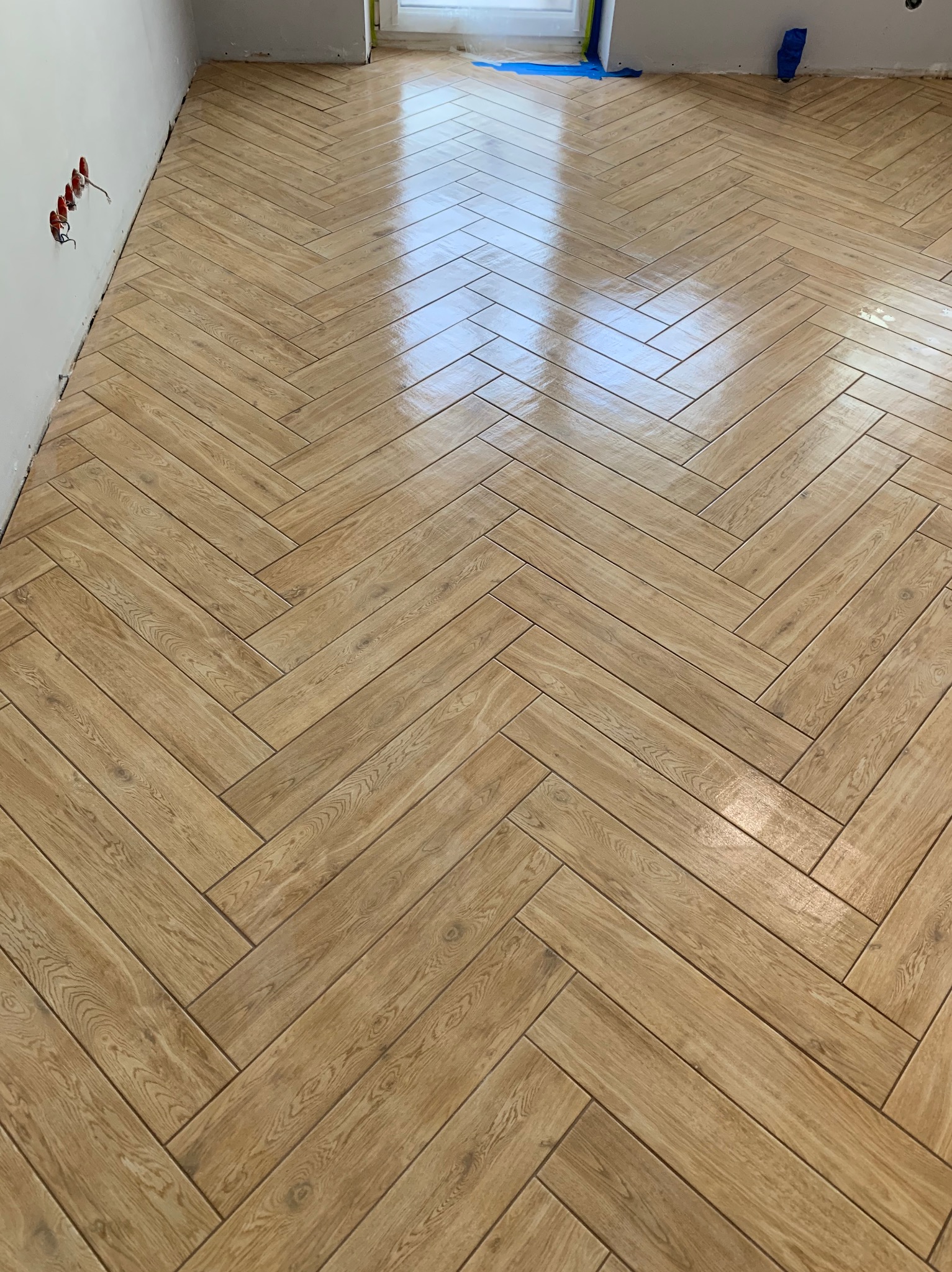 Ułożona, błyszcząca podłoga z płytek imitujących drewno w jodełkę, widoczne gniazdka elektryczne i zabezpieczenia folią malarską przy ścianie.