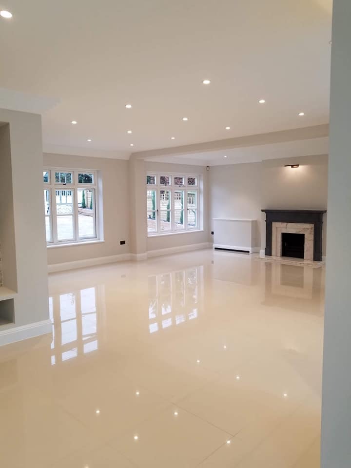 Jasna, przestronna sala z błyszczącą, beżową podłogą z płytek, białymi ścianami, oknami z widokiem na zieleń i czarnym kominkiem z marmurowym obramowaniem.