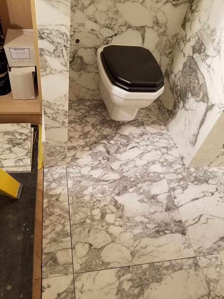 Łazienka wykończona płytkami imitującymi marmur, z widoczną toaletą z czarną deską oraz fragmentem regału z pudełkami i płytkami, ukazująca etap prac wykończeniowych.