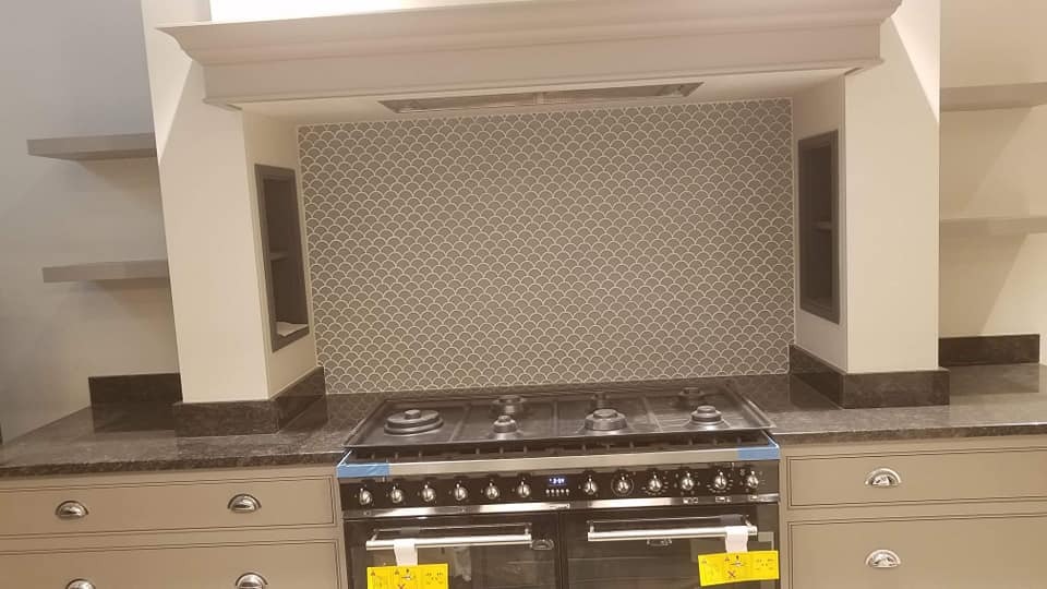 Wykończona kuchnia z szarą mozaiką w kształcie łusek nad kuchenką, zabudowa w odcieniach szarości z ciemnym blatem, widoczne półki i zabudowany okap.