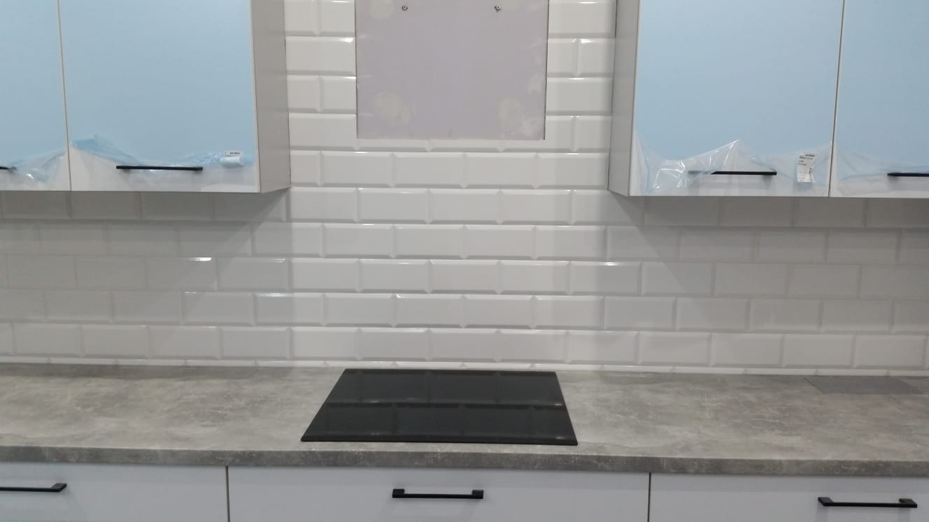 Nowoczesna kuchnia z białymi, błyszczącymi płytkami typu metro, szarym blatem i jasnoniebieskimi szafkami, niektóre jeszcze zabezpieczone folią ochronną, z widoczną płytą indukcyjną i przygotowanym...