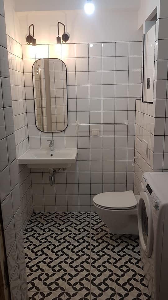 Biała łazienka z czarno-białą mozaikową podłogą, wyposażona w umywalkę, lustro z zaokrąglonymi rogami, toaletę i pralkę.