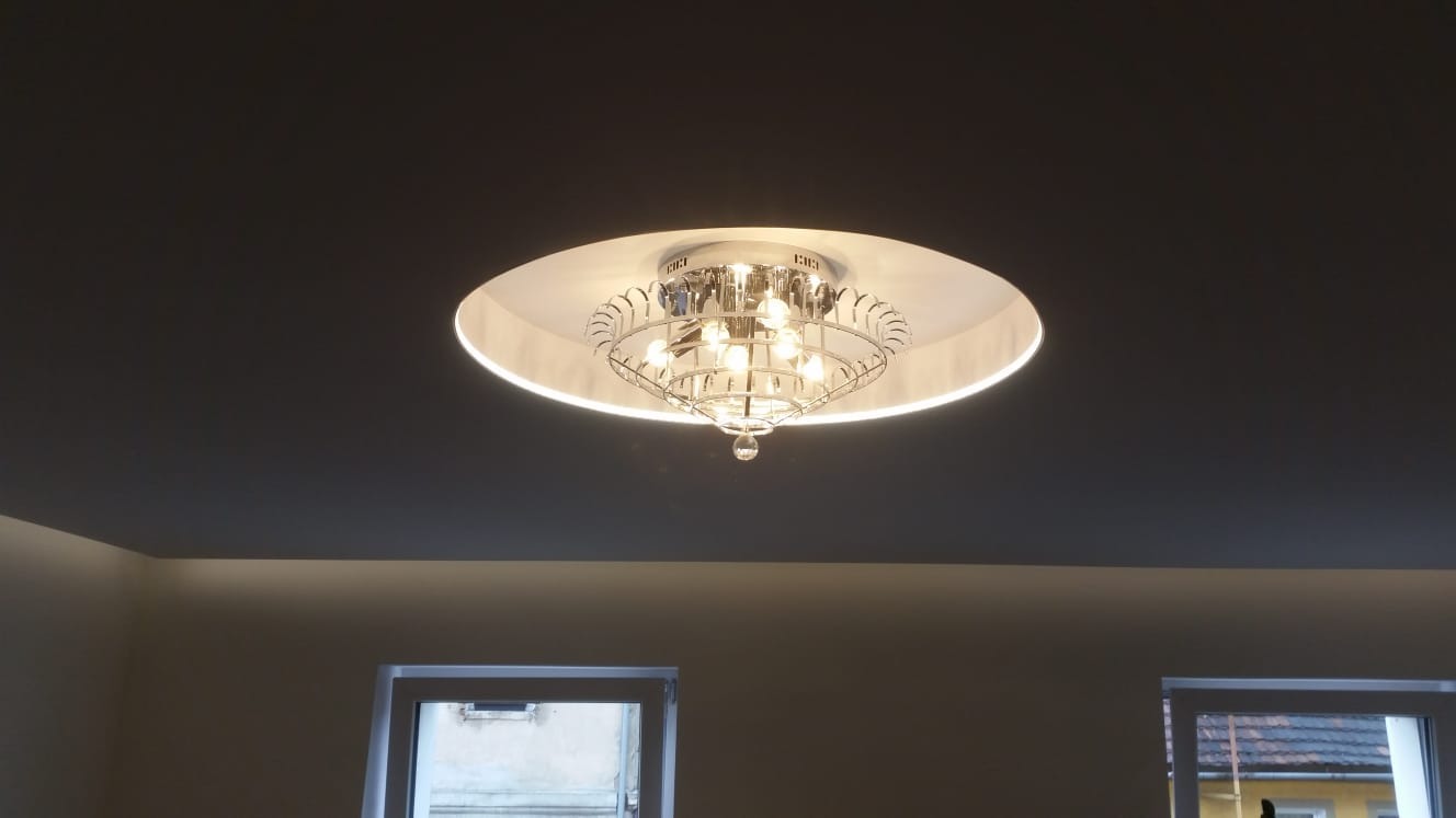 Kryształowy żyrandol z okrągłą obudową zamontowany na ciemnoszarym suficie w pomieszczeniu z dwoma oknami.