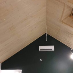 GERARD-PRO USŁUGI RMONTOWO-BUDOWLANE MICHAŁ BZICKI - Skośny sufit wykończony boazerią w jasnym odcieniu drewna, kontrastujący z ciemnozieloną ścianą, na której zamontowano klimatyzator i widoczne są wystające kable oświetleniowe.