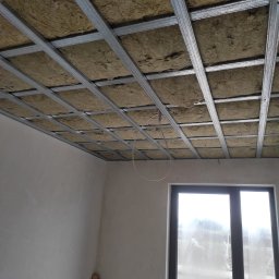 GERARD-PRO USŁUGI RMONTOWO-BUDOWLANE MICHAŁ BZICKI - Sufit podwieszany z metalową konstrukcją wypełnioną wełną mineralną, widok z dołu na ocieplone poddasze w trakcie prac wykończeniowych, okno w tle.
