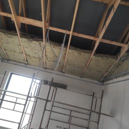 GERARD-PRO USŁUGI RMONTOWO-BUDOWLANE MICHAŁ BZICKI - Montaż izolacji z wełny mineralnej na poddaszu, widoczne drewniane krokwie, czarna membrana dachowa, przewody elektryczne oraz rusztowanie.