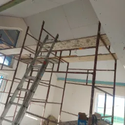 Montaż płyt na skośnym poddaszu, widoczne rusztowanie, drabina i częściowo zamontowane płyty z widocznymi wkrętami.