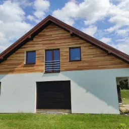 Dwukondygnacyjny budynek z elewacją w kolorze jasnego drewna na górze i białym tynkiem na dole, z brązowym dachem i trzema oknami, z widocznymi otworami wejściowymi i częściowo opuszczoną roletą...