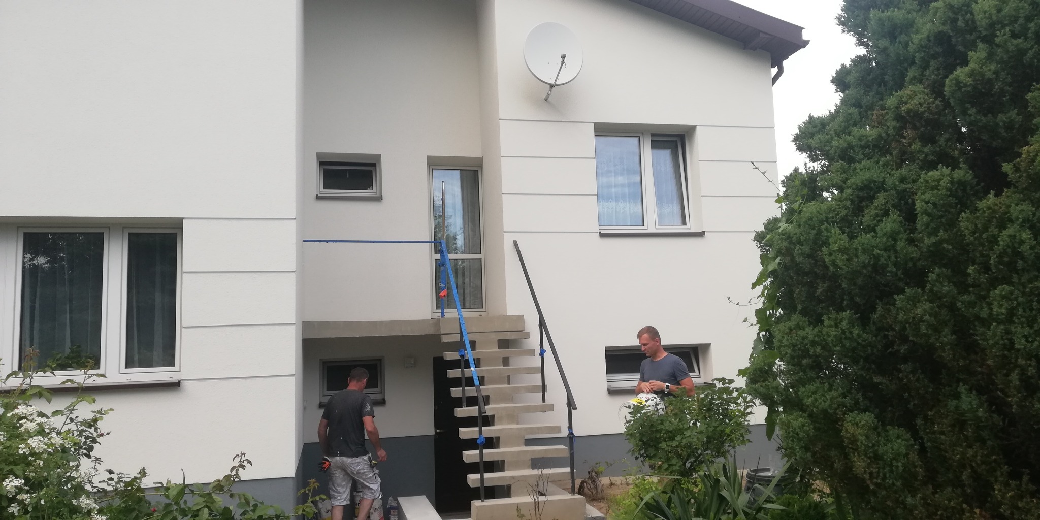 Dwóch pracowników w Ciecierzynie przygotowuje się do malowania elewacji dwupiętrowego domu z betonowymi schodami wejściowymi, widoczna satelita na ścianie.