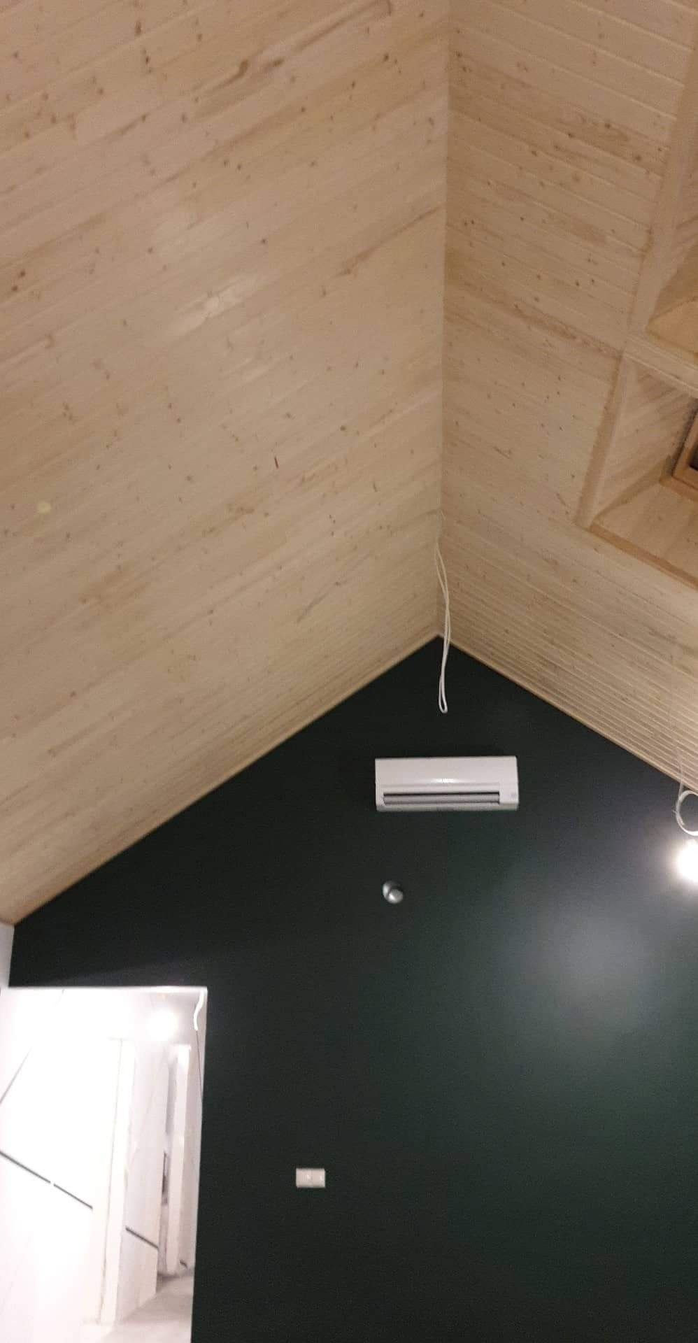Skośny sufit wykończony boazerią w jasnym odcieniu drewna, kontrastujący z ciemnozieloną ścianą, na której zamontowano klimatyzator i widoczne są wystające kable oświetleniowe.