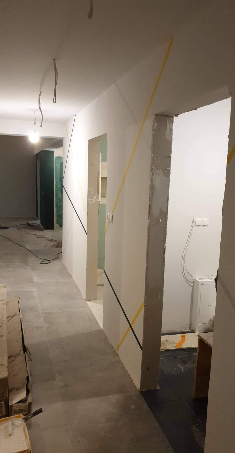 Korytarz w trakcie wykańczania z geometrycznymi wzorami malarskimi, widoczne taśmy malarskie w kolorze żółtym i czarnym, surowy beton na podłodze i brakujące ościeżnice.