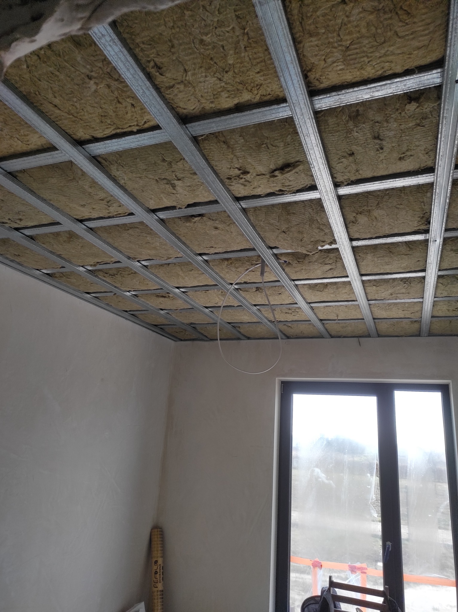 Sufit podwieszany z metalową konstrukcją wypełnioną wełną mineralną, widok z dołu na ocieplone poddasze w trakcie prac wykończeniowych, okno w tle.