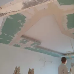 Montaż sufitu podwieszanego z płyt kartonowo-gipsowych, widoczne profile, szpachlowanie łączeń, drabina drewniana i pracownik z kielnią.