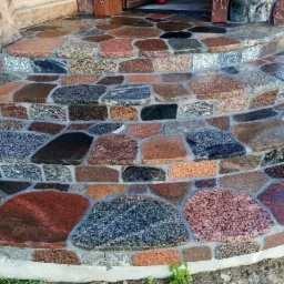 Schody wejściowe wyłożone nieregularnymi kamieniami z granitu w odcieniach szarości, czerwieni i brązu, z fugami wypełnionymi zaprawą, prowadzące do drewnianego wejścia.