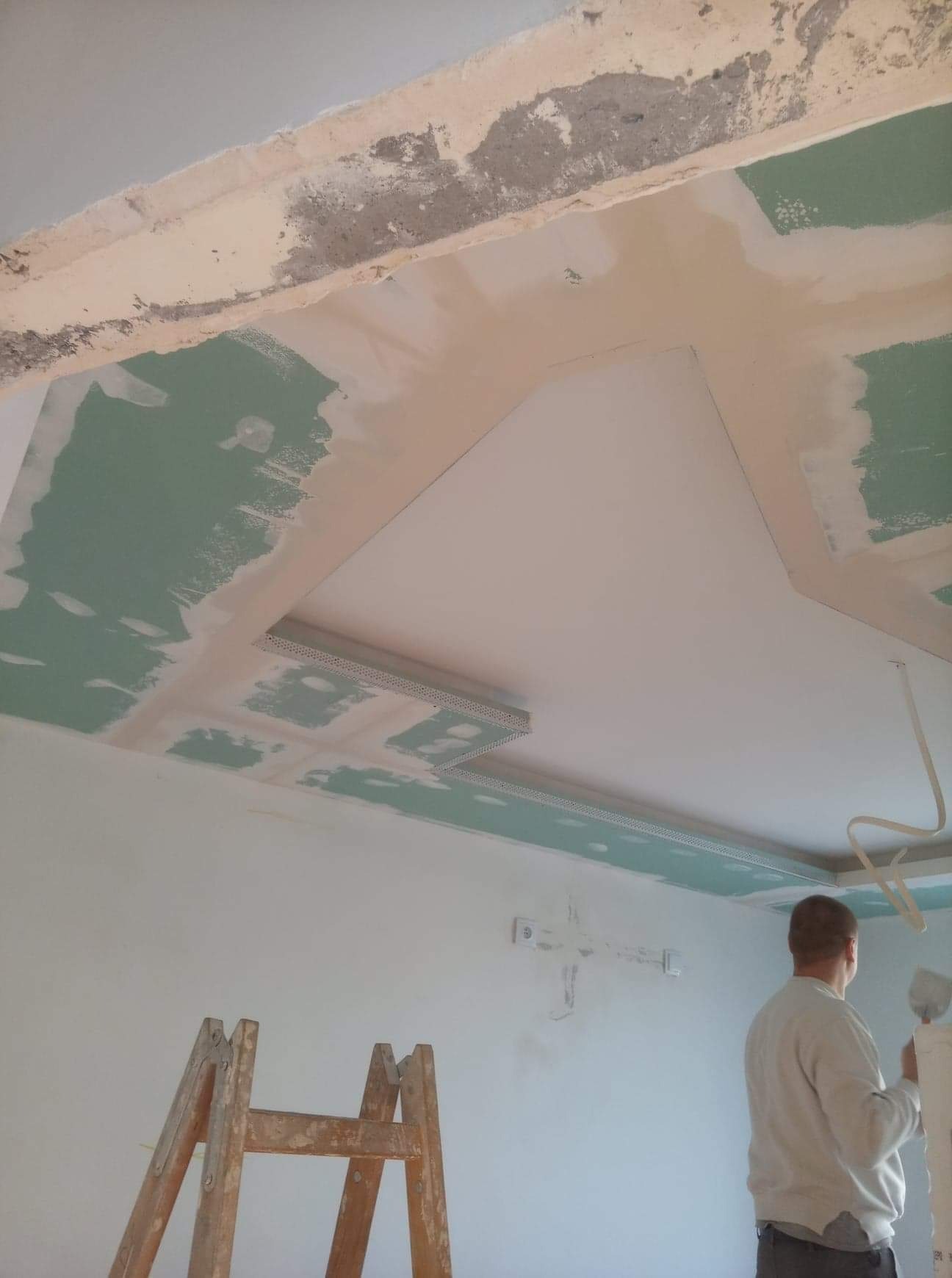 Montaż sufitu podwieszanego z płyt kartonowo-gipsowych, widoczne profile, szpachlowanie łączeń, drabina drewniana i pracownik z kielnią.