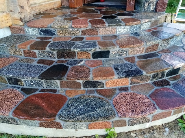 Schody wejściowe wyłożone nieregularnymi kamieniami z granitu w odcieniach szarości, czerwieni i brązu, z fugami wypełnionymi zaprawą, prowadzące do drewnianego wejścia.