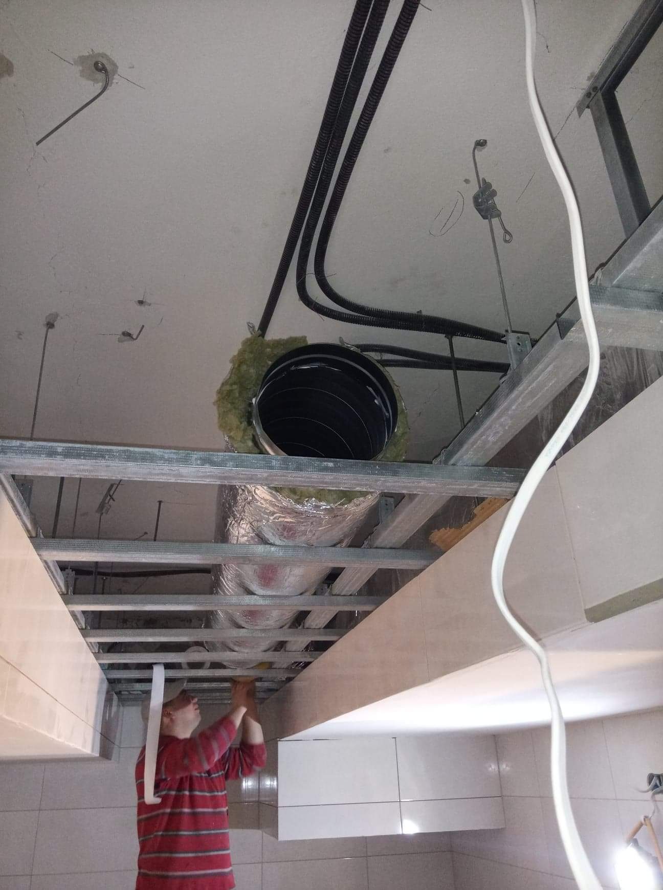 Montaż sufitu podwieszanego z widoczną konstrukcją metalową, izolacją i instalacją wentylacyjną w Grudziądzu, wykonywany przez instalatora w czerwonej koszuli.