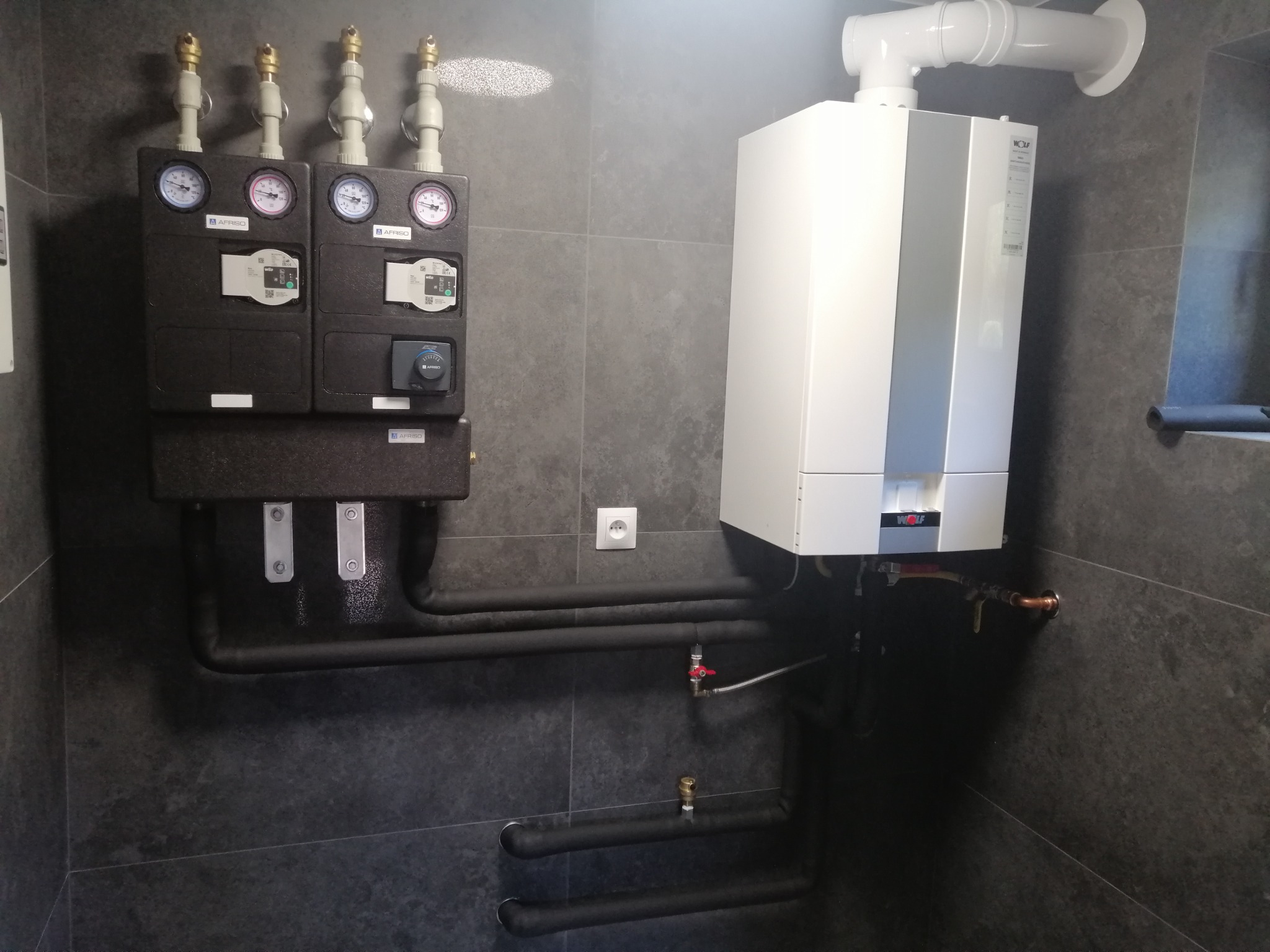 Nowoczesny, biały piec centralnego ogrzewania marki Wolf zawieszony na szarej, płytek ceramicznych ścianie, z widocznymi czarnymi rurami instalacji grzewczej i czarną skrzynką z manometrami...