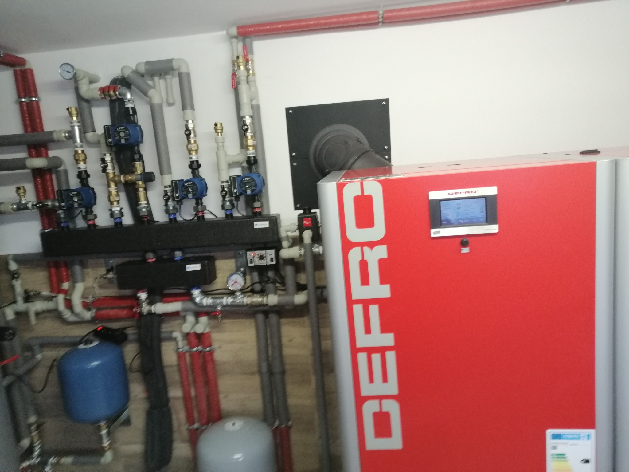 Czerwony piec CO marki DEFRO z podłączoną instalacją grzewczą, widoczne rury, pompy obiegowe i zbiornik wyrównawczy, całość zamontowana na ścianie.