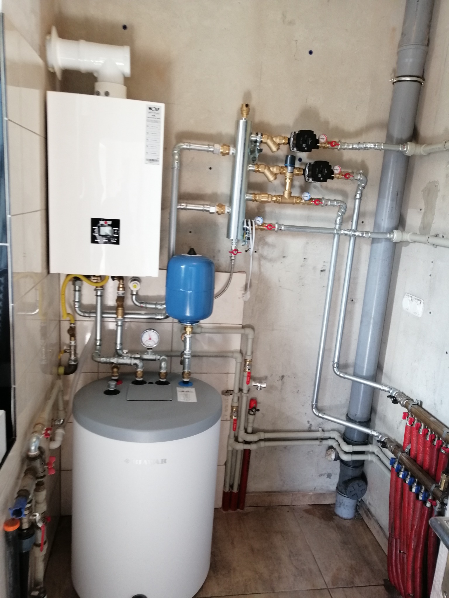 Biały piec gazowy i zasobnik ciepłej wody z niebieskim naczyniem przeponowym, liczne rury instalacji CO i CWU z zaworami, manometrami i pompami obiegowymi, szara rura kanalizacyjna w rogu...