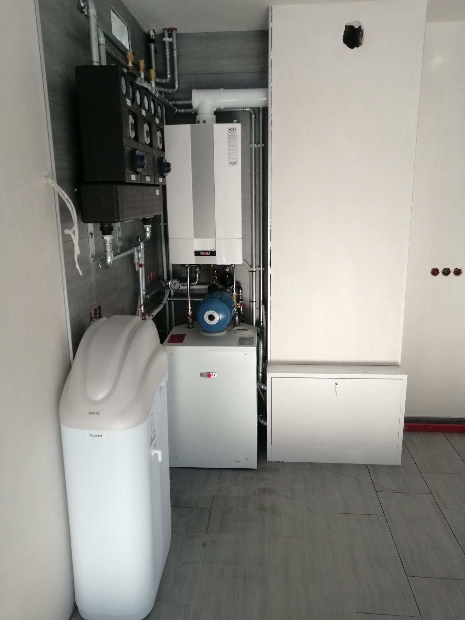 Nowoczesna kotłownia: biały piec gazowy Wolf, czarna armatura, niebieska pompa obiegowa, filtr do wody Primo, szare płytki podłogowe, białe ściany z widocznymi otworami instalacyjnymi.
