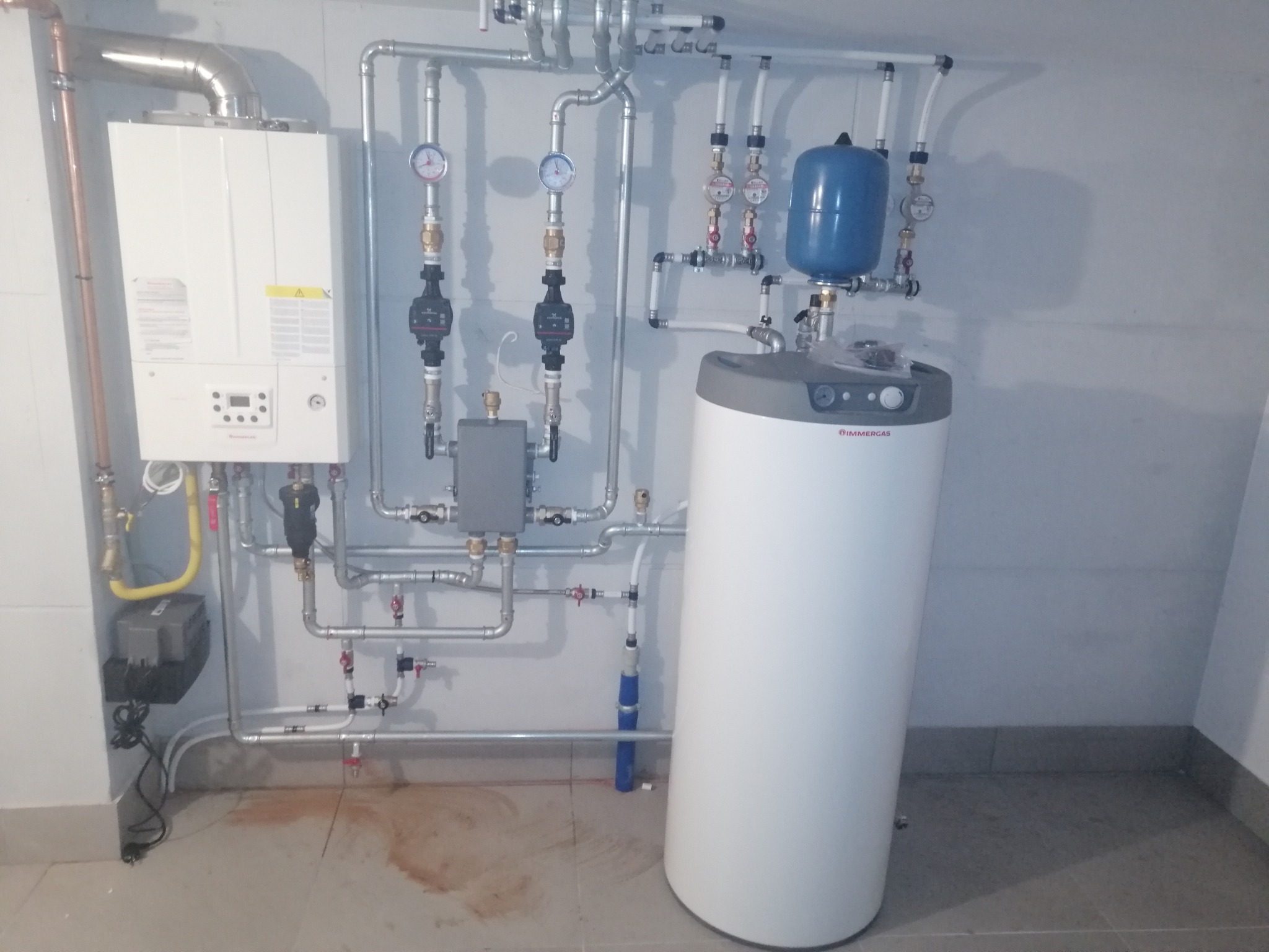 Zainstalowany, wiszący piec gazowy Immergas z odprowadzeniem spalin, obok biały zasobnik ciepłej wody z niebieskim naczyniem wzbiorczym i rozbudowaną instalacją hydrauliczną z zaworami, manometrami...