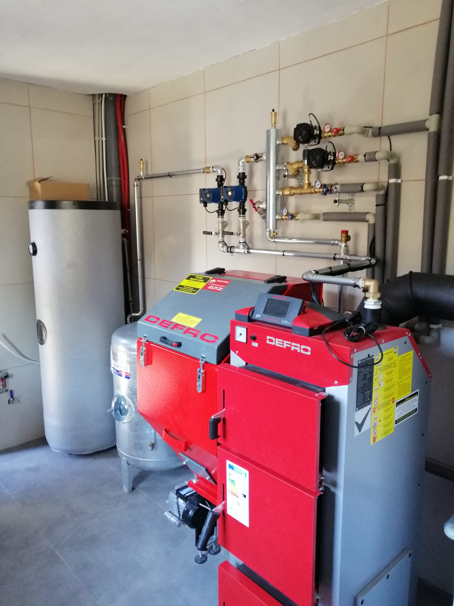 Kotłownia z czerwono-szarym piecem Defro, zasobnikiem wody i instalacją grzewczą z rurami w izolacji, widok na kompletną instalację w pomieszczeniu z beżowymi płytkami.