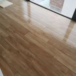 Ułożone pod kątem brązowe płytki podłogowe imitujące drewno, z widocznym odbiciem światła, przy dużym oknie z widokiem na ceglany budynek.