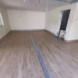 Układanie podłogi z paneli imitujących drewno w jasnym, przestronnym pokoju z odpływem liniowym; widoczne niedokończone prace wykończeniowe ścian i sufitu.