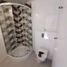 Nowoczesna łazienka z kabiną prysznicową z wzorzystymi płytkami w odcieniach szarości i brązu, podwieszaną toaletą, na której leży wkrętarka akumulatorowa i instrukcja montażu.