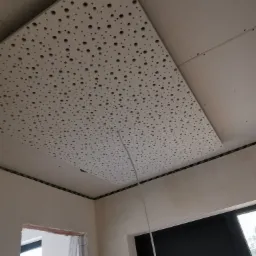 Montaż perforowanej płyty karton-gips na suficie, widoczne kable i fragment drabiny w tle, pomieszczenie w trakcie remontu.