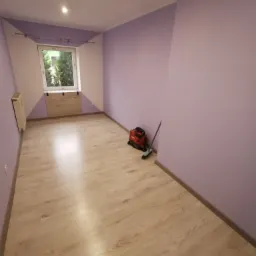 Pomalowany na liliowo pokój z jasnymi panelami podłogowymi, widoczny grzejnik, zakryte okno i odkurzacz Hilti z miotłą przy ścianie.