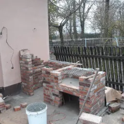 Budowa murowanego grilla z czerwonej cegły na zewnątrz, widoczne zapasy cegieł, wiadro z zaprawą, drewniany płot w tle.