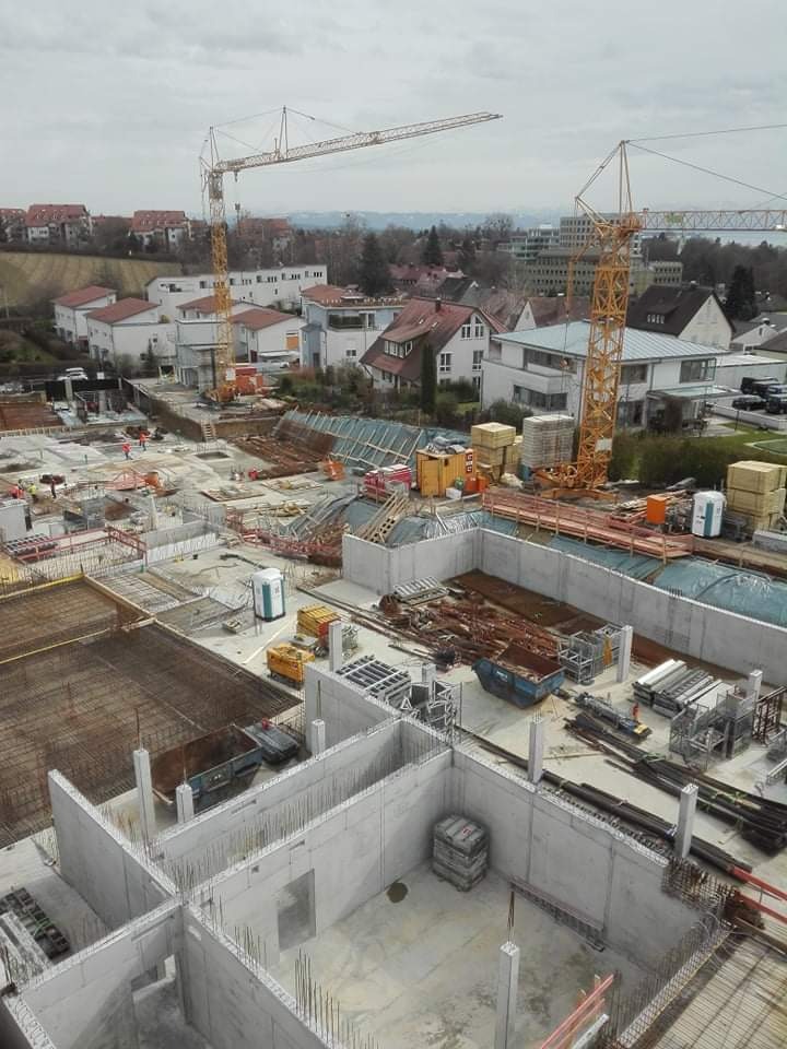 Budowa domu murowanego: widok z góry na fundamenty i ściany parteru w trakcie wznoszenia, z dwoma żurawiami wieżowymi w tle i zabudową mieszkalną w oddali.