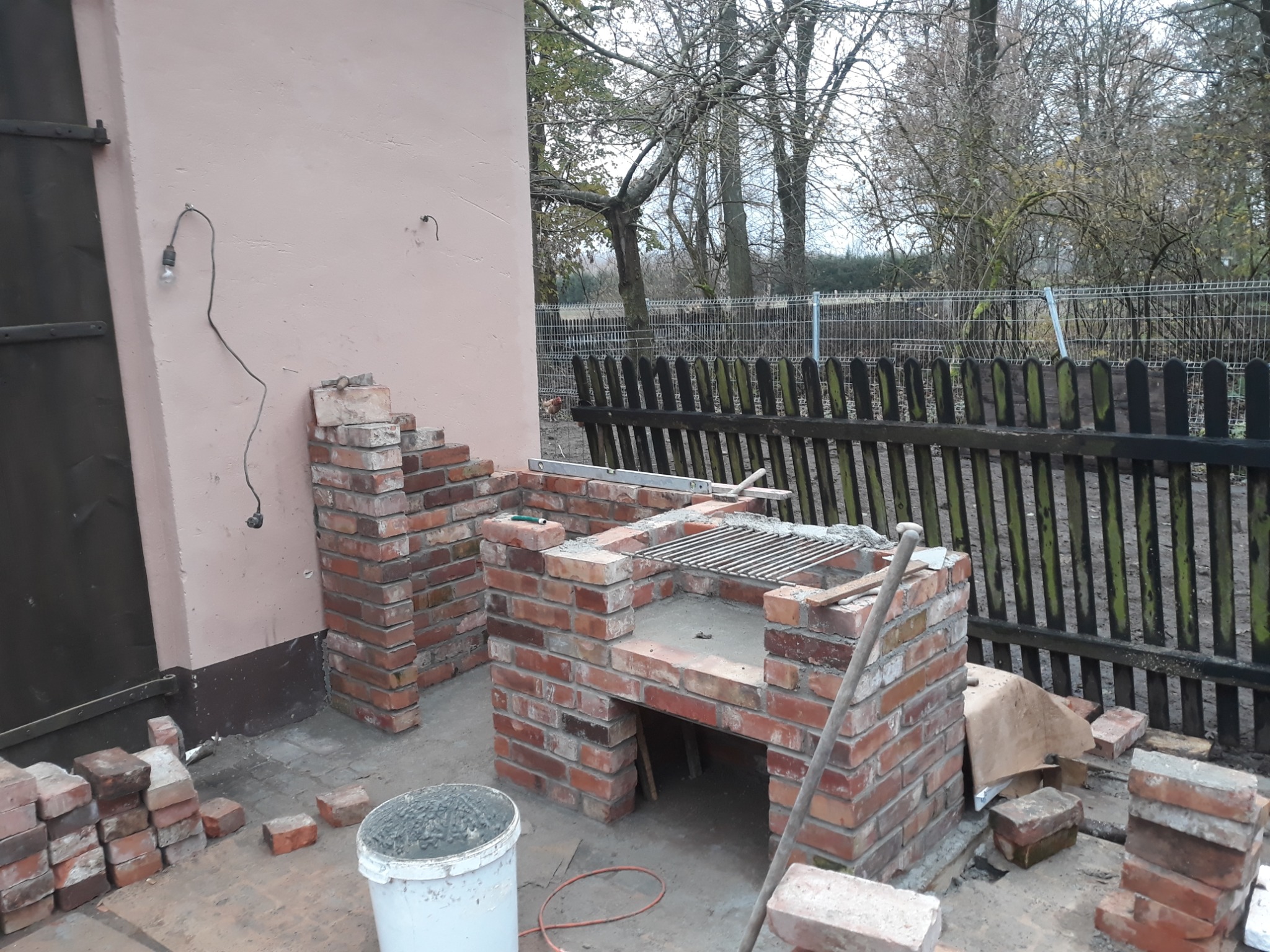 Budowa murowanego grilla z czerwonej cegły na zewnątrz, widoczne zapasy cegieł, wiadro z zaprawą, drewniany płot w tle.