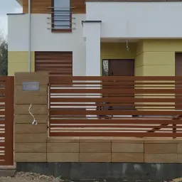 Nowoczesne ogrodzenie z poziomych, brązowych profili stalowych i słupk&oacute;w z piaskowca, z wideodomofonem i skrzynką na listy, na tle domu z ż&oacute;łtymi akcentami.