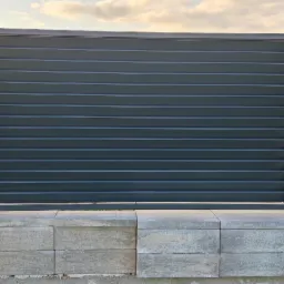 Nowoczesna brama wjazdowa z poziomych, szarych profili metalowych, osadzona w słupkach z szarej kostki betonowej o zróżnicowanej fakturze. Widoczne elementy mocujące. Słupki zróżnicowane...