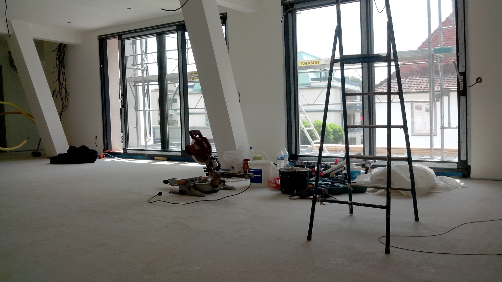 Wnętrze w trakcie prac wykończeniowych z widoczną piłą ukosową, drabiną i narzędziami na podłodze, duże okna z widokiem na rusztowanie za oknem.