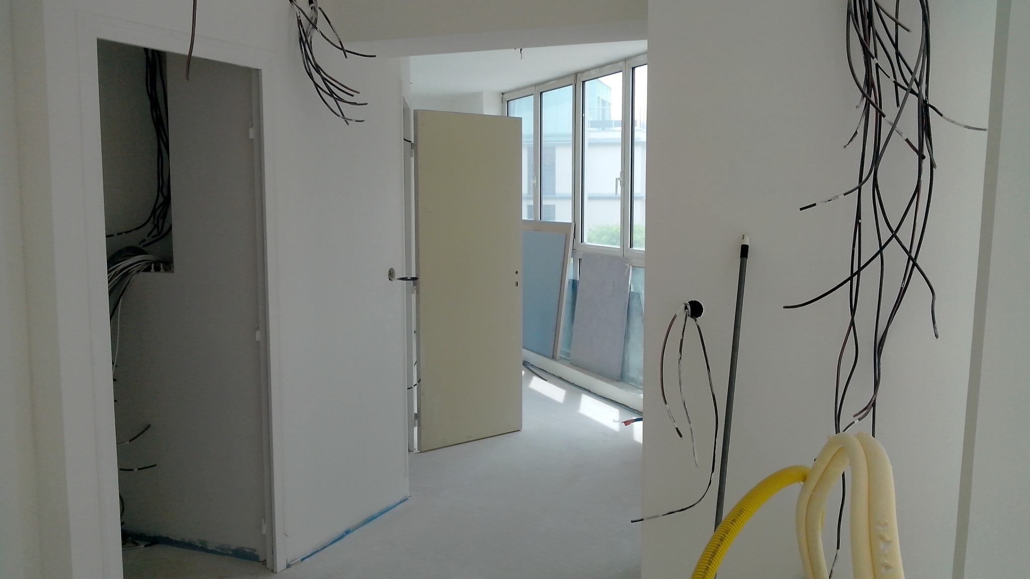 Wnętrze w trakcie remontu: widoczne kable elektryczne zwisające ze ścian, rury wentylacyjne, surowe ściany przygotowane do wykończenia, drzwi wewnętrzne z widoczną instalacją elektryczną wewnątrz...