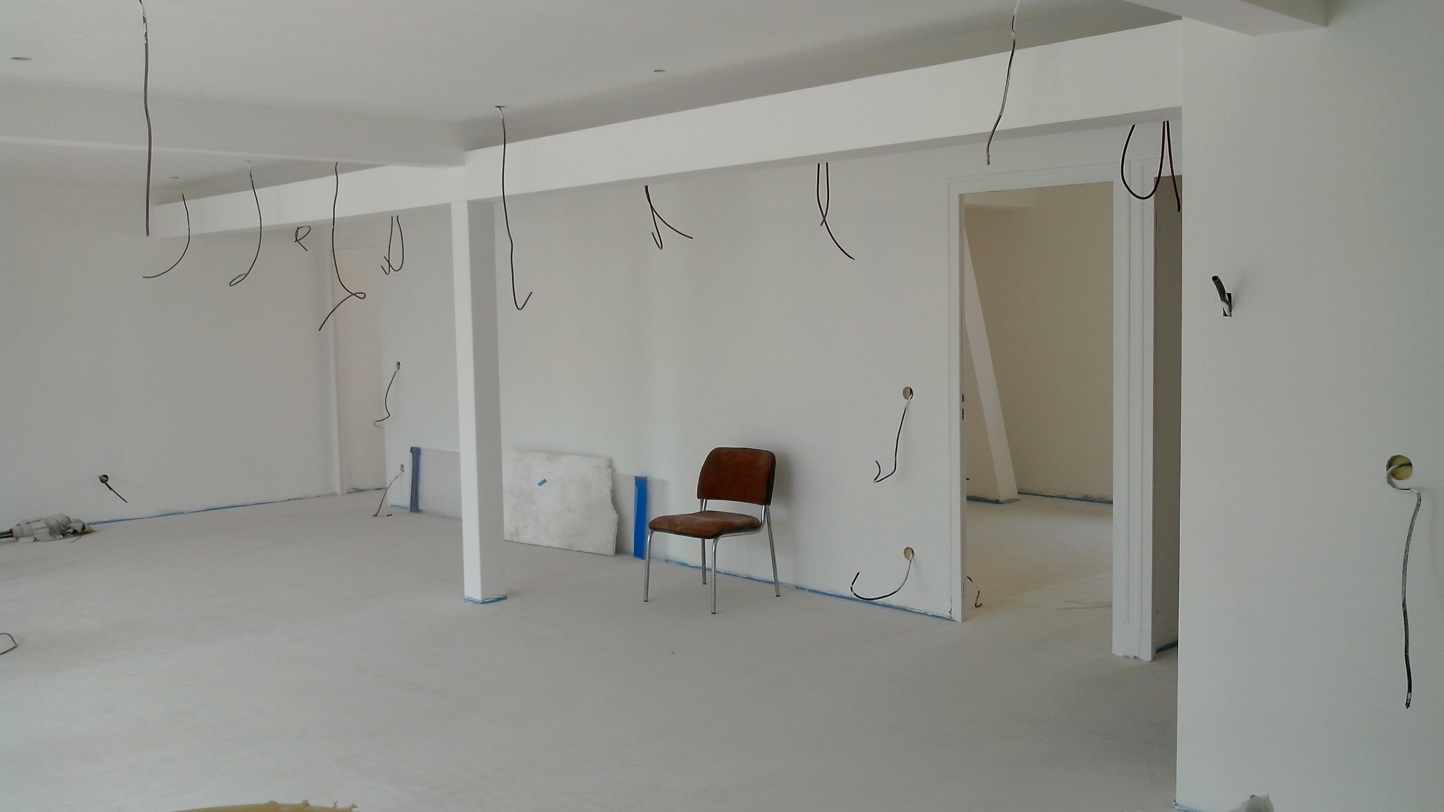 Wnętrze w trakcie remontu: białe ściany, surowa podłoga, wystające przewody elektryczne i samotne krzesło na środku pomieszczenia.