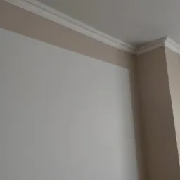 Pomieszczenie po remoncie: ściana w kolorze złamanej bieli z beżowym pasem pod sufitem i białym, ozdobnym gzymsem.