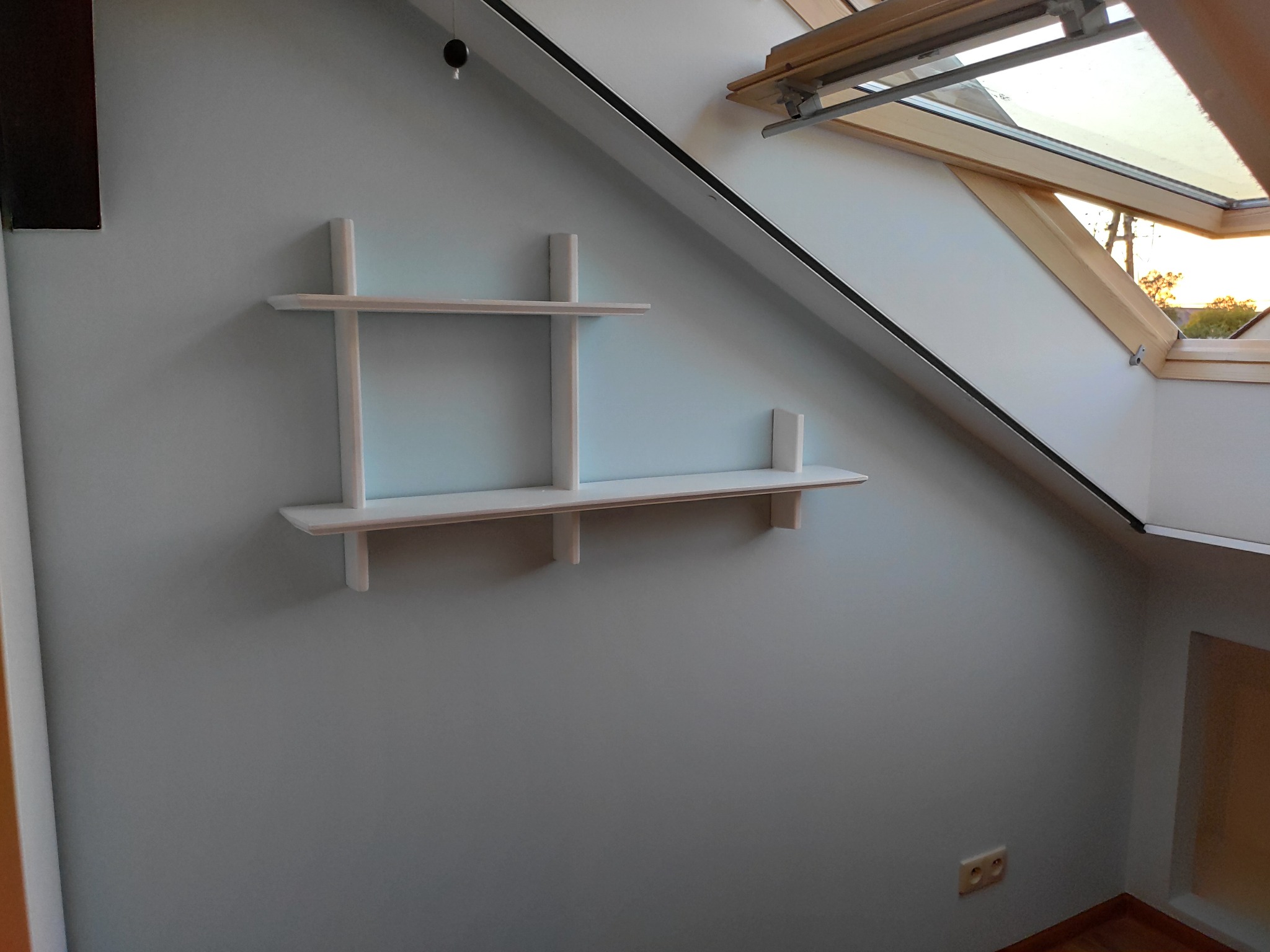 Ściana w pokoju na poddaszu po odświeżeniu, z zamontowanymi białymi półkami i oknem dachowym, widoczny gładki, jednolity kolor szaro-błękitny.