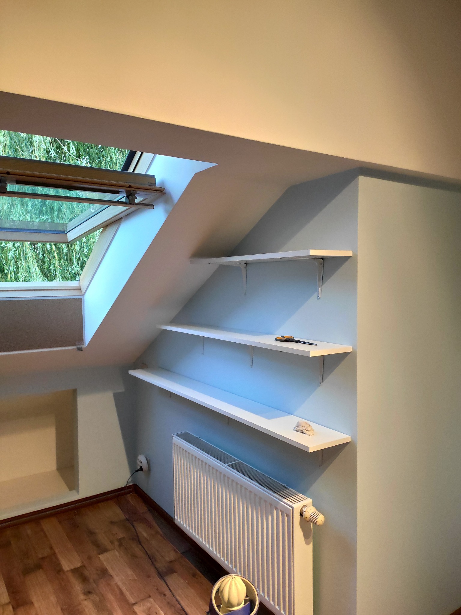 Pokój na poddaszu po odświeżeniu ścian w odcieniach błękitu, z zamontowanymi białymi półkami i grzejnikiem pod oknem dachowym.